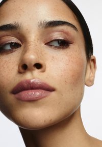 Gros plan sur un visage avec un teint lisse, de légères taches de rousseur, des lèvres pleines avec du rouge à lèvres mauve et un maquillage des yeux doux et naturel.