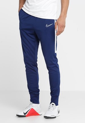 Man die marineblauwe Nike trainingsbroek met witte streep en witte sportschoenen met rode en zwarte accenten draagt, staand tegen een effen achtergrond.