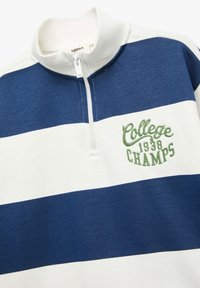 Pusero, jossa on valkoisia ja laivaston sinisiä raitoja, neljännesvetoketju kauluksessa ja vihreät brodeeraukset "College 1938 CHAMPS". Pehmeää puuvillamateriaalia.