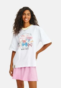 Biela grafická tričko s viacfarebnými mašľami a textom, vo voľnom strihu, skombinované s ružovou sukňou. Má krátke rukávy a štítok s dizajnom.