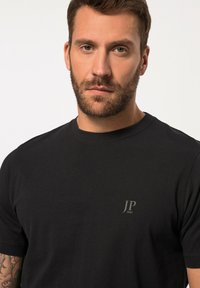 Svart t-shirt i bomull med rund hals, korta ärmar och en liten "JP 1880"-logotyp på vänster bröst. Mjuk textur.