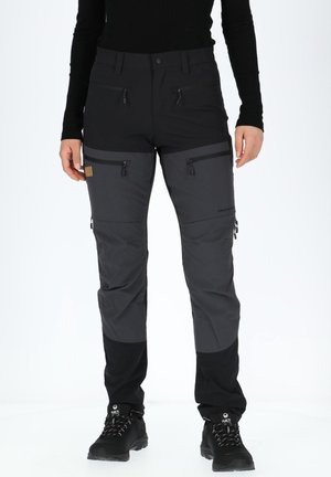 Personne portant un pantalon d'extérieur noir et gris foncé avec plusieurs poches zippées et des chaussures de randonnée noires.