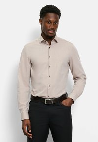 Langärmeliges, hellbeiges Hemd mit feiner Struktur, bis zum Kragen zugeknöpft. Verfügt über dunkle Knöpfe und eine schlanke Passform. Kombiniert mit dunkler Hose und Gürtel.