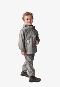 Graue wasserdichte Jacke mit einer Fronttasche, elastischen Bündchen und passenden Hosen. Mit einem sternförmigen Etikett versehen. Braune Stiefel runden das Outfit ab.
