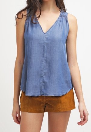 Ärmelloses blaues Denim-V-Ausschnitt-Oberteil kombiniert mit braunen Wildleder-Shorts, getragen von einer stehenden Person vor einfarbigem Hintergrund.
