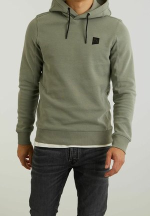 Homme portant un sweat à capuche vert clair avec cordons noirs, superposé sur une chemise blanche, associé à un jean gris foncé.