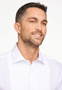 Camicia bianca con texture frontale, colletto a punta e chiusura a bottoni, presentata su uno sfondo neutro con un modello sorridente.