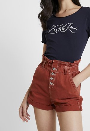 Kobieta ubrana w dopasowany granatowy t-shirt z białym napisem oraz wysokim stanem rdzawoczerwone szorty z podwiniętymi mankietami i guzikami.
