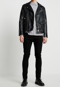 Osoba ubrana w czarną skórzaną kurtkę motocyklową, jasnoszary zapinany na guziki strój, czarne wąskie spodnie i czarne sznurowane buty stoi na tle jednolitej ściany.