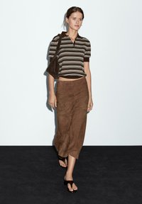 Brunstribet kortærmet polo skjorte med lynlås, parret med en lang brun ruskinds nederdel. Modellen bærer sorte flade sandaler og har en matchende taske.