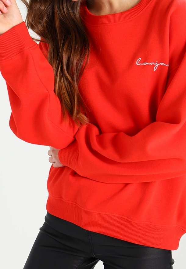 Femme portant un sweat-shirt rouge vif avec un texte blanc en cursive sur la poitrine, associé à un pantalon noir, bras croisés à la taille.