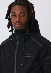 Giacca nera impermeabile con zip frontale, cappuccio regolabile con cordino e dettagli riflettenti. Presenta il logo sul petto e una tasca laterale con zip.