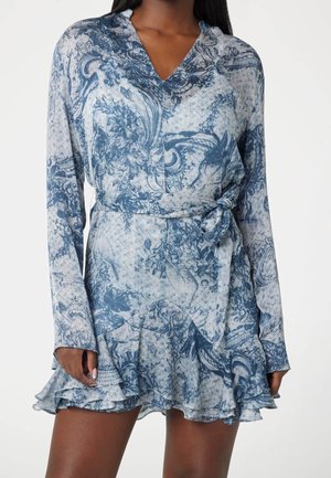 Kobieta ubrana w niebiesko-białą sukienkę kopertową w kwiatowy wzór z długimi rękawami i falbaną przy dole na białym tle.