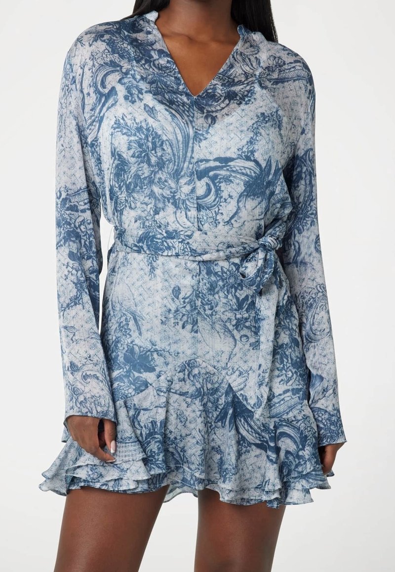 Femme portant une robe portefeuille à manches longues avec un motif floral bleu et blanc et un ourlet à volants sur fond blanc.
