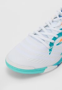 Chaussure de sport blanche avec des accents turquoise, dessus en maille texturée, motifs de rayures diagonales et lacets plats. Semelle en caoutchouc conçue pour l'adhérence.
