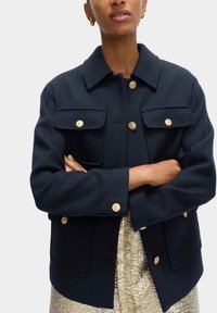 Marineblaue Wolljacke mit Kragen, Goldknopfverschlüssen, zwei Brusttaschen und lockerer Passform, kombiniert mit einem strukturierten goldenen Rock.