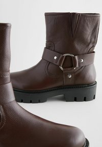 Botas de tobillo de cuero marrón con acabado texturizado, que cuentan con una correa ancha y un detalle de anillo de metal, y una suela gruesa de goma negra.