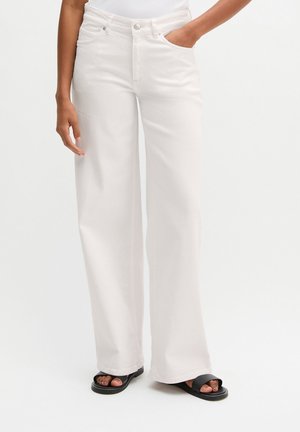 Osoba nosząca białe spodnie z wysokim stanem i szerokimi nogawkami z przednimi kieszeniami, w połączeniu z czarnymi sandałami z odkrytymi palcami na jednolitym białym tle.