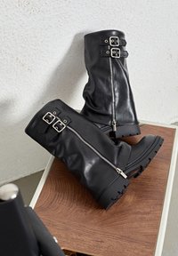 Bottes en cuir noir hautes jusqu'au genou avec une fermeture éclair et deux accents de boucle en haut. La semelle en caoutchouc solide offre une bonne traction.