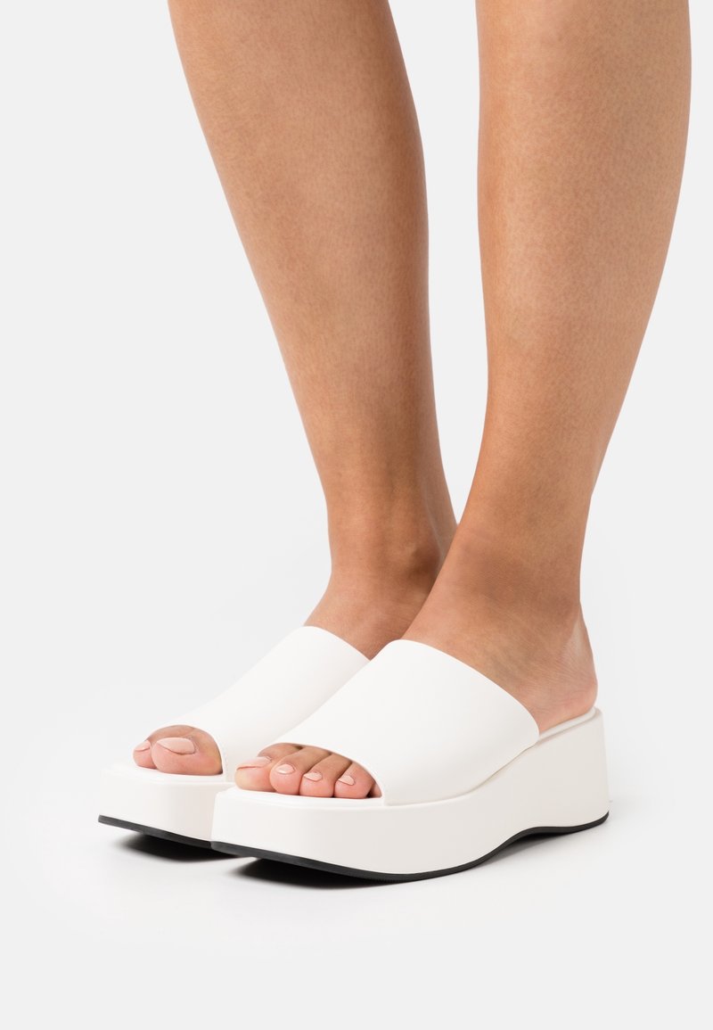 Pieds portant des sandales blanches à plateforme avec orteils ouverts et semelles noires sur fond blanc.