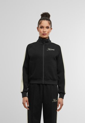 Giacca nera con zip e dettagli a strisce crema sulle maniche, abbinata a pantaloni jogger. Presenta un colletto alto e un logo ricamato. Tessuto liscio.