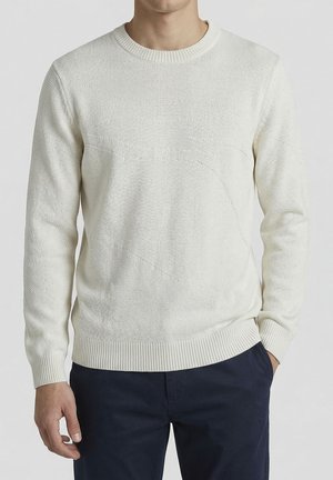 Uomo che indossa un maglione bianco in maglia testurizzata con polsini e orlo a coste, abbinato a pantaloni blu scuro, in piedi contro uno sfondo neutro.