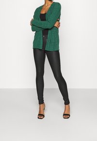 Zielony kardigan z otwartym przodem, wykonany z teksturowanej dzianiny, noszony na czarnej bluzce i dopasowanych czarnych spodniach, w połączeniu z czarnymi sandałkami na obcasie.