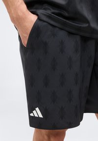 Άνδρας με μαύρο σορτς με σχέδιο και λογότυπο Adidas, χέρι που ακουμπά στην πλαϊνή τσέπη, φαίνεται από τη μέση μέχρι το μέσο του μηρού.