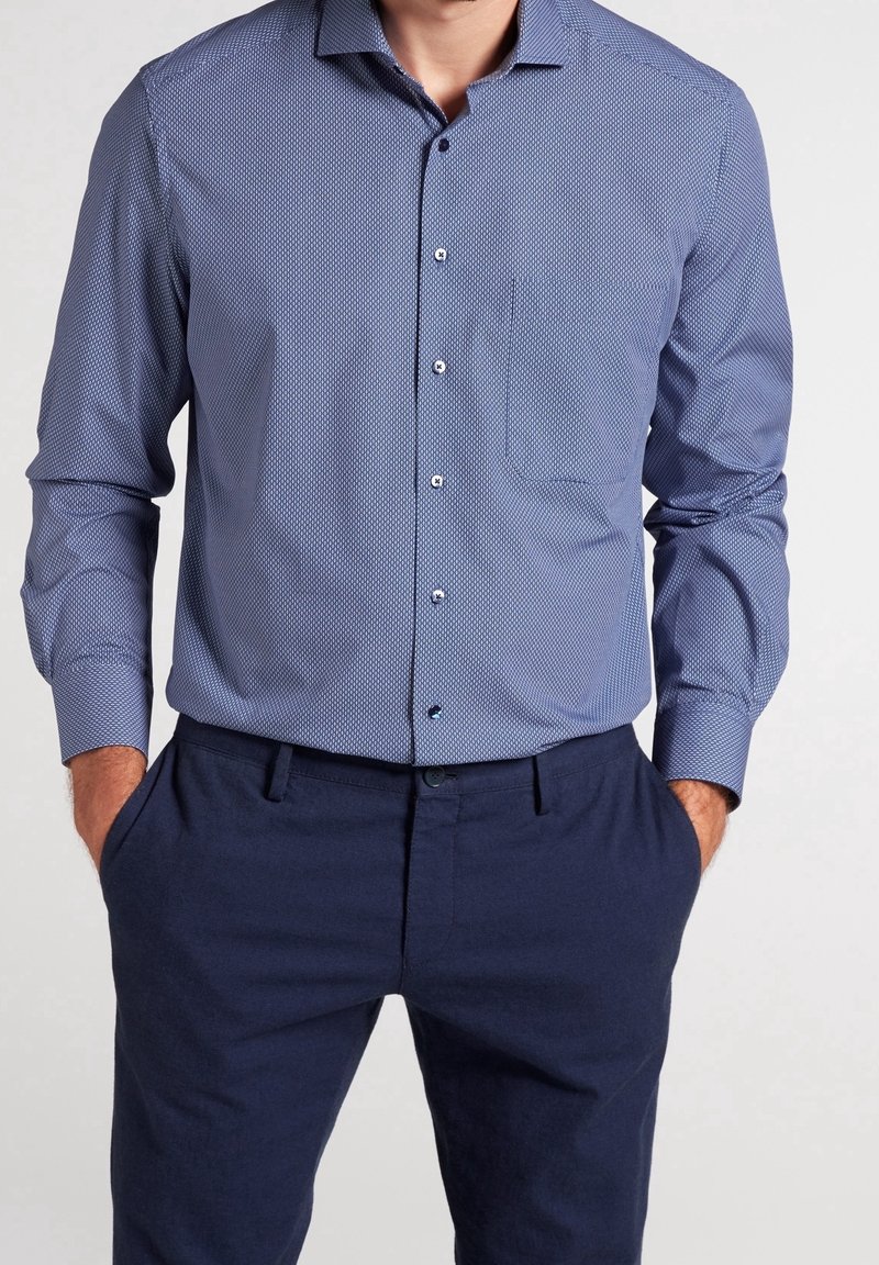Homme portant une chemise de ville ajustée à manches longues bleue à motifs, rentrée dans un pantalon bleu foncé, les mains dans les poches.