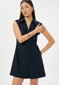 Robe courte marine avec un col, un devant boutonné et des manches courtes. Dispose d'une taille cintrée et d'une jupe évasée.