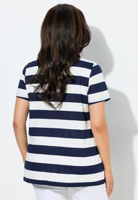 Frau mit langen, welligen, dunklen Haaren, die ein marineblau-weiß gestreiftes, kurzärmeliges Oberteil und weiße Hosen trägt, steht vor einer weißen Wand.