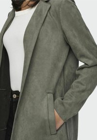 Kurze grüne Wildlederjacke mit Reverskragen, seitlichen Taschen und glatter Textur, getragen über einem weißen Oberteil.