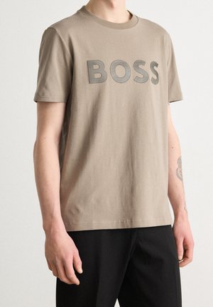 Beige short-sleeve t-shirt with large "BOSS" text across the chest, worn with black pants, shown from neck to hips.
