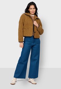 Hnědá fleecová bunda s krátkým střihem, kontrastními detaily, předními kapsami, nošená přes béžový top a v kombinaci s modrými širokými džíny.