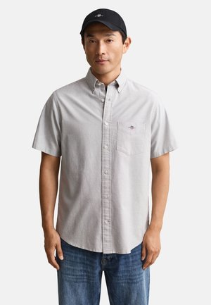 Mężczyzna w jasnoszarej, krótkiej koszuli zapinanej na guziki z logo na kieszeni, niebieskich dżinsach i czarnej czapce z daszkiem, stojący na jednolitym tle.