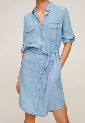 Robe chemise en denim bleu clair avec manches longues retroussées, boutonnière devant, deux poches poitrine et ceinture nouée à la taille.