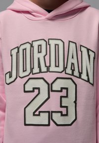 Rosa hettegenser med stor hvit tekst "JORDAN" og "23" foran, med mørke konturer. Mykt stoff med standard pullover-design.