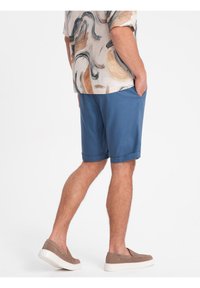 Shorts bleus en tissu léger, avec un ourlet replié et des poches latérales. Une chemise à motifs à manches courtes est portée au-dessus.