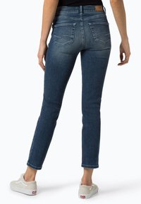Jean skinny bleu foncé à taille mi-haute, poches arrière avec détails de coutures, et texture douce en denim. Associé à des baskets de couleur claire.