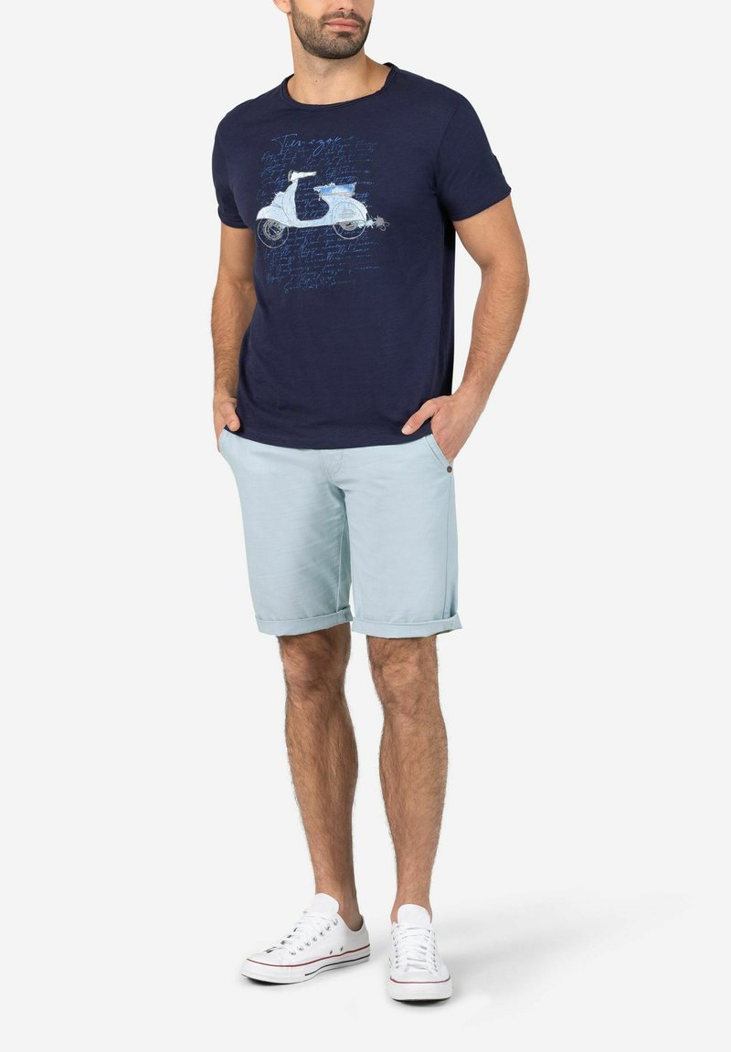 Marineblaues T-Shirt mit einem Scooter-Graphic und Text, kombiniert mit hellblauen, hochgekrempelten Shorts und weißen Turnschuhen. Material aus Baumwollmischung.