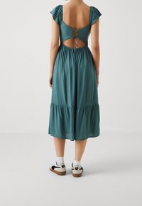 Tyrkysové midi šaty s nafouknutými rukávy, otevřeným zadním dílem a šněrovacím detailem, s nařaseným páskem a stupňovitým lemem. Nosené s bílými teniskami a ponožkami.