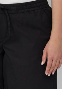 Rechte Hand ruht neben einer schwarzen Hose mit elastischem Bund und vorderen Taschen, trägt ein weißes Oberteil.