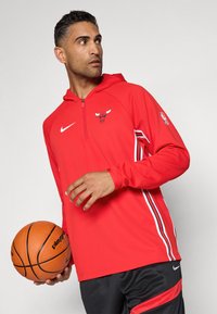 Rød Nike hettegenser med halv glidelås, med Bulls-logo, hvite sidelister og svarte treningsshorts. Hånd holder en basketball.