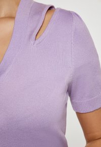 Top scurt, tricotat, de culoare violet deschis, cu decolteu în V și detalii decupate pe umeri. Textură netedă, formă ajustată și manșete cu model în relief.