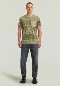 Khaki camouflage T-shirt met een gestructureerd patroon en een borstzak, gecombineerd met blauwe jeans en zwarte laarzen. Model staat rechtop.