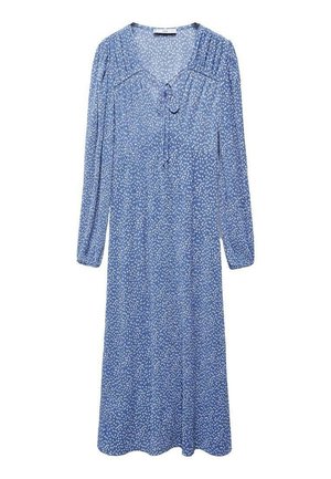 Frau trägt ein blaues Midikleid mit weißen Sprenkeln, langen gerafften Ärmeln, V-Ausschnitt und goldenen flachen Schuhen und steht vor einem schlichten Hintergrund.