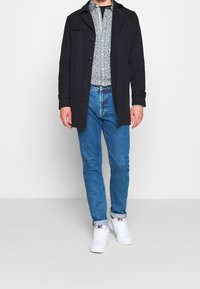 Svart ullblandad kappa med knäppning, mönstrad skjorta under, ljusblå denimjeans och vita sneakers med färgglada detaljer.