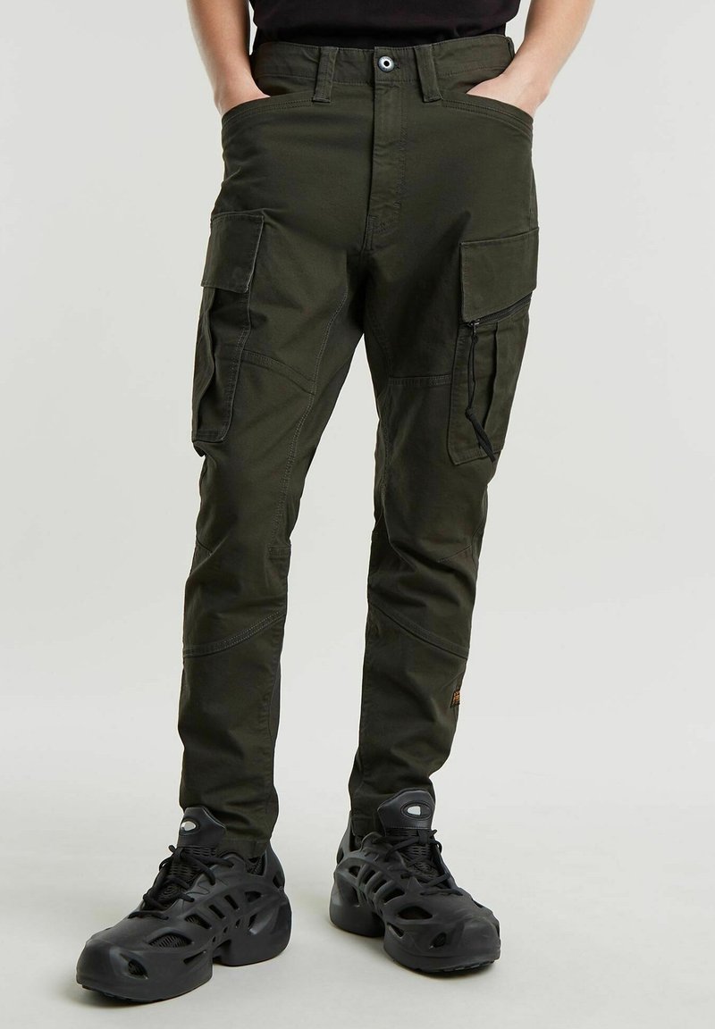 Homme portant un pantalon cargo vert foncé avec plusieurs poches et des baskets noires épaisses, debout les mains dans les poches.