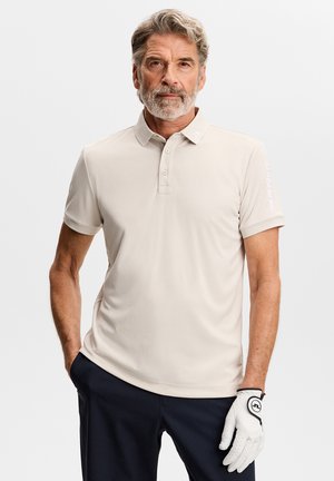 Uomo di mezza età con capelli grigi e barba che indossa un polo beige a maniche corte e pantaloni neri, posando con la mano destra in tasca e un guanto bianco sulla sinistra.