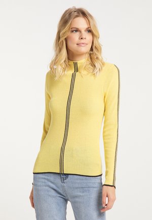 Lyshåret kvinde iført en ribbet gul langærmet sweater med sorte lodrette striber og lyseblå jeans mod en ensfarvet baggrund.